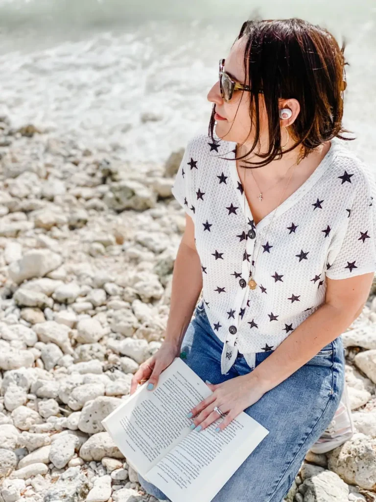 Francesca reading a paperback with an earbud in, listening and reading by the water.