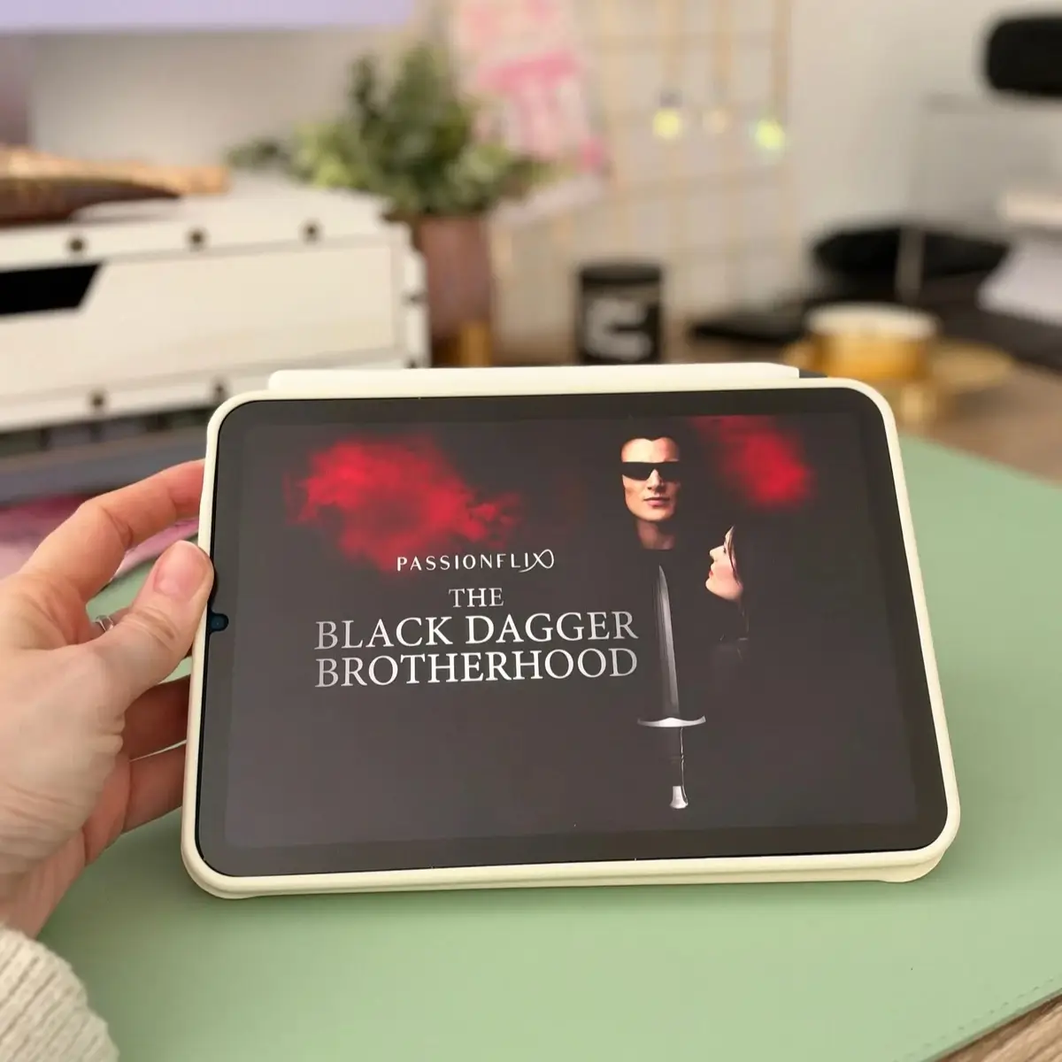 A hand holding an iPad showing the Passionflix title screen for The Black Dagger Brotherhood TV show.