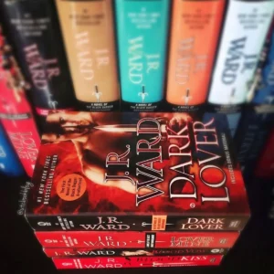 A paperback of Dark Lover by J.R. Ward on top of a stack with Lover Mine, Blood Vow and Blood Kiss, in front of a row of J.R. Ward hardcovers in red, gold, teal, orange and white