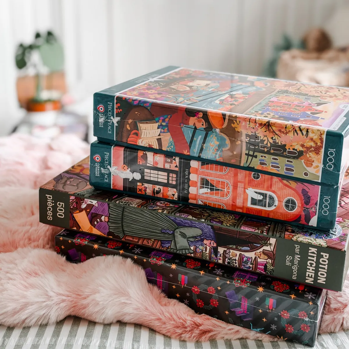 A stack of four boxed jigsaw puzzles on a pink faux fur throw: Autumn in Amsterdam, Trick or Treat, Potion Kitchen and the Midnight Glow advent calendar