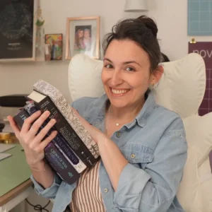 Francesca smiling in a white chair, hugging a stack of J.R. Ward books including two copies of the Black Dagger Brotherhood 20th Anniversary Insider's Guide, Lover Enthroned and Crown of War and Shadow