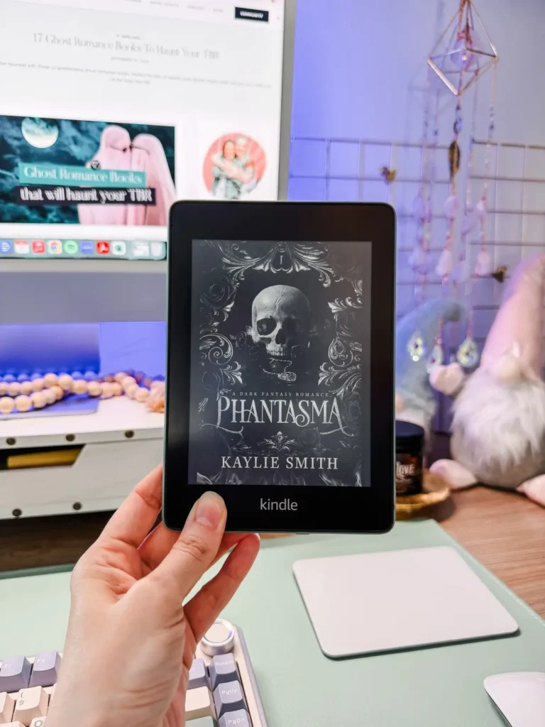 Hand holding a Kindle with the cover of Phantasma by Kaylie Smith on screen, in front of Francesca's desk with her monitor and a plush gnome