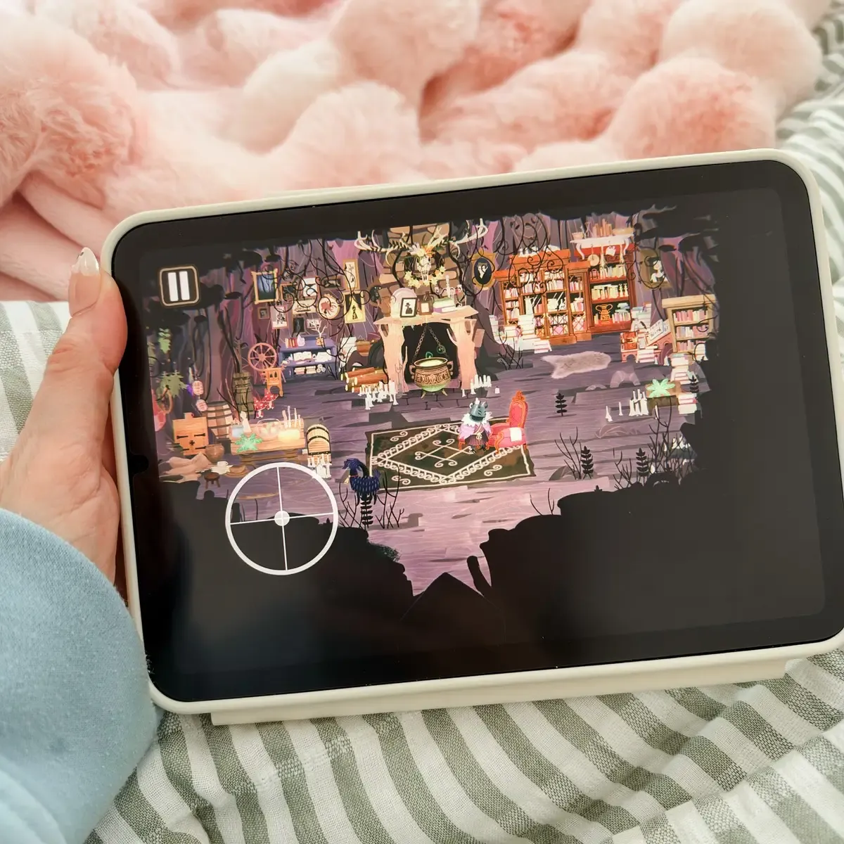 Holding an iPad playing Wytchwood on a striped blanket, the witch sitting in her cluttered cottage beside a bubbling cauldron, bookshelves and candles